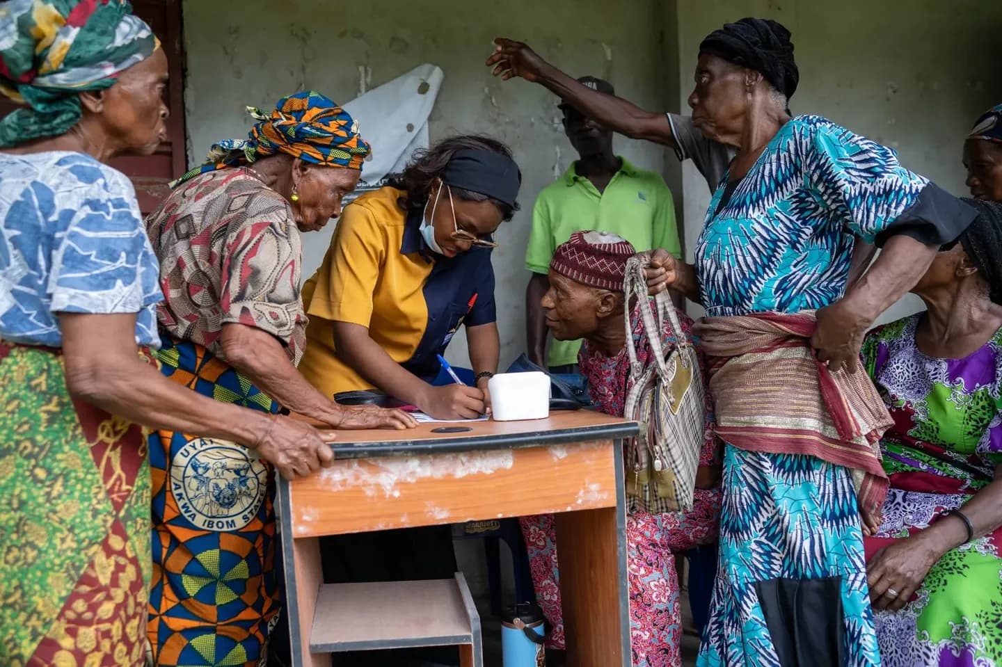 A GAMAID volunteer writes down details at a small desk as elderly villagers gather round to register