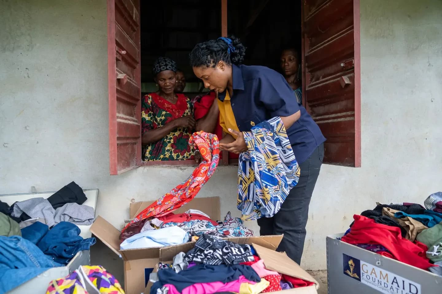 A GAMAID volunteer picks out clothing from boxes of donated items while women look on from a window