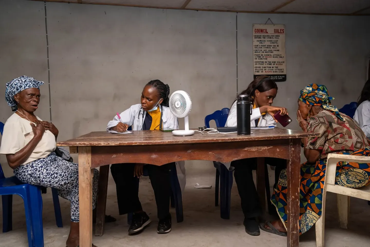 Two medical volunteers in white coats attend to two elderly women at a wooden table in the village council hall