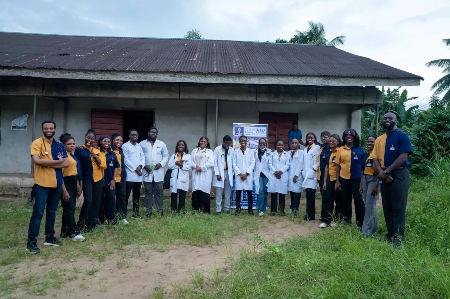 The GAMAID outreach team, volunteers in gold and navy shirts and medical personnel in white coats, standing outside the hall at Atan Ekpe