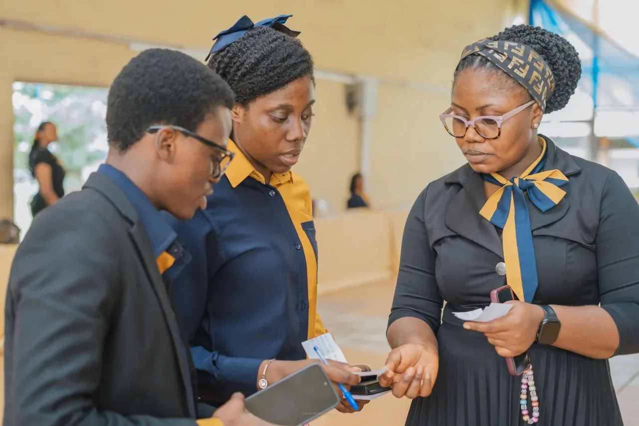Three GAMAID volunteers in navy and gold confer over notes at an event